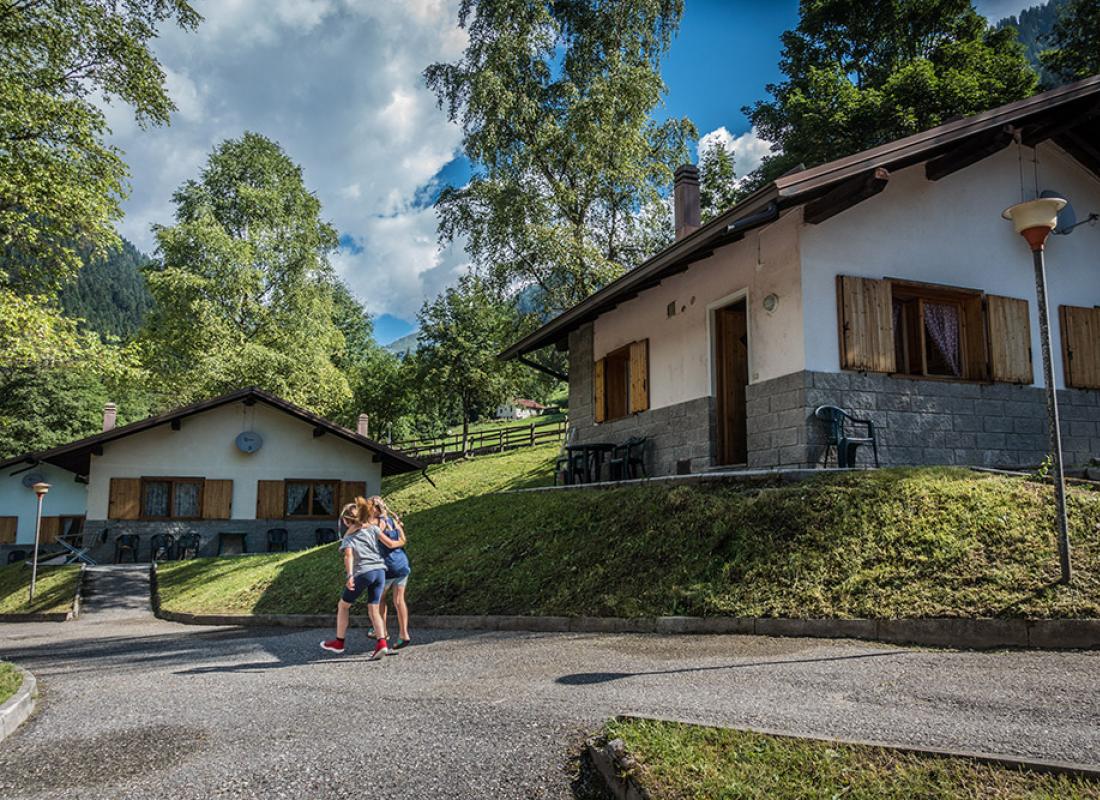 Due persone camminano davanti a case in montagna, circondate da alberi e natura.