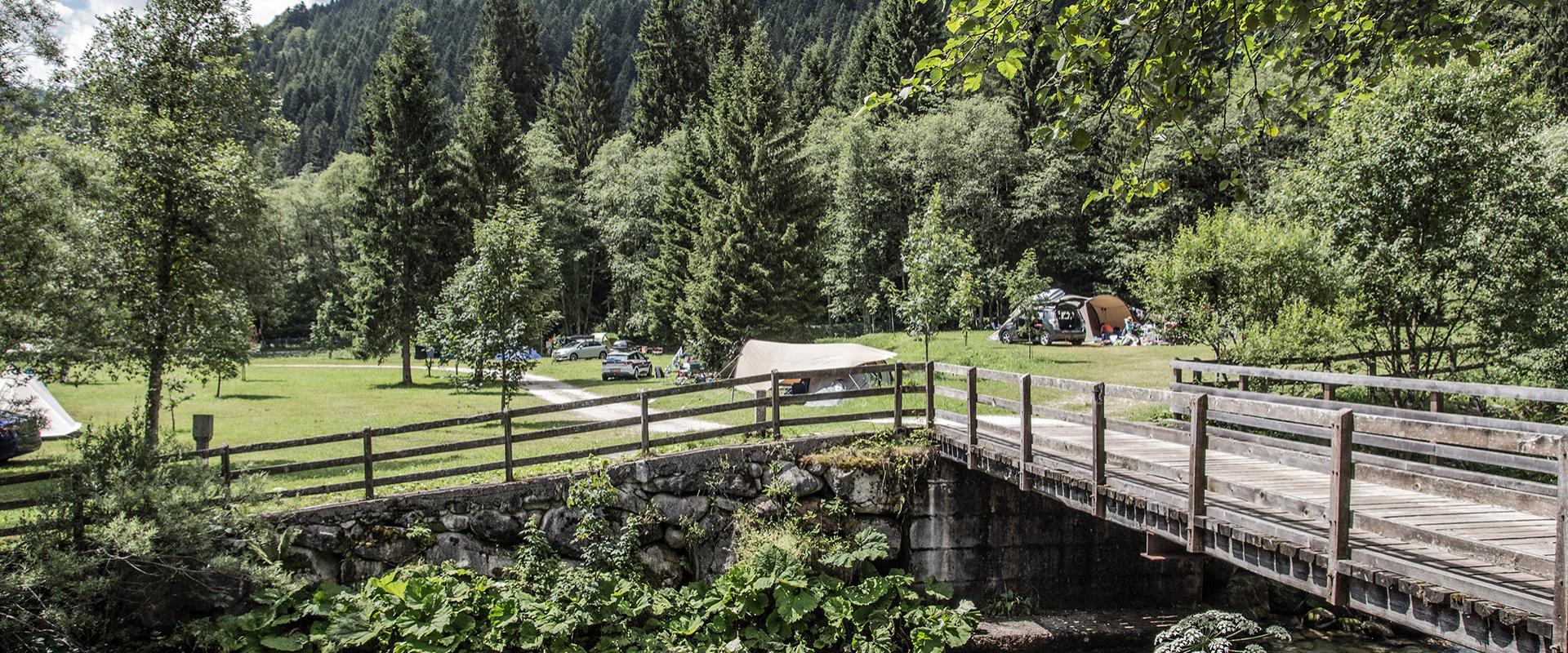 Paesaggio montano con ponte di legno, fiume e area campeggio immersa nella natura.