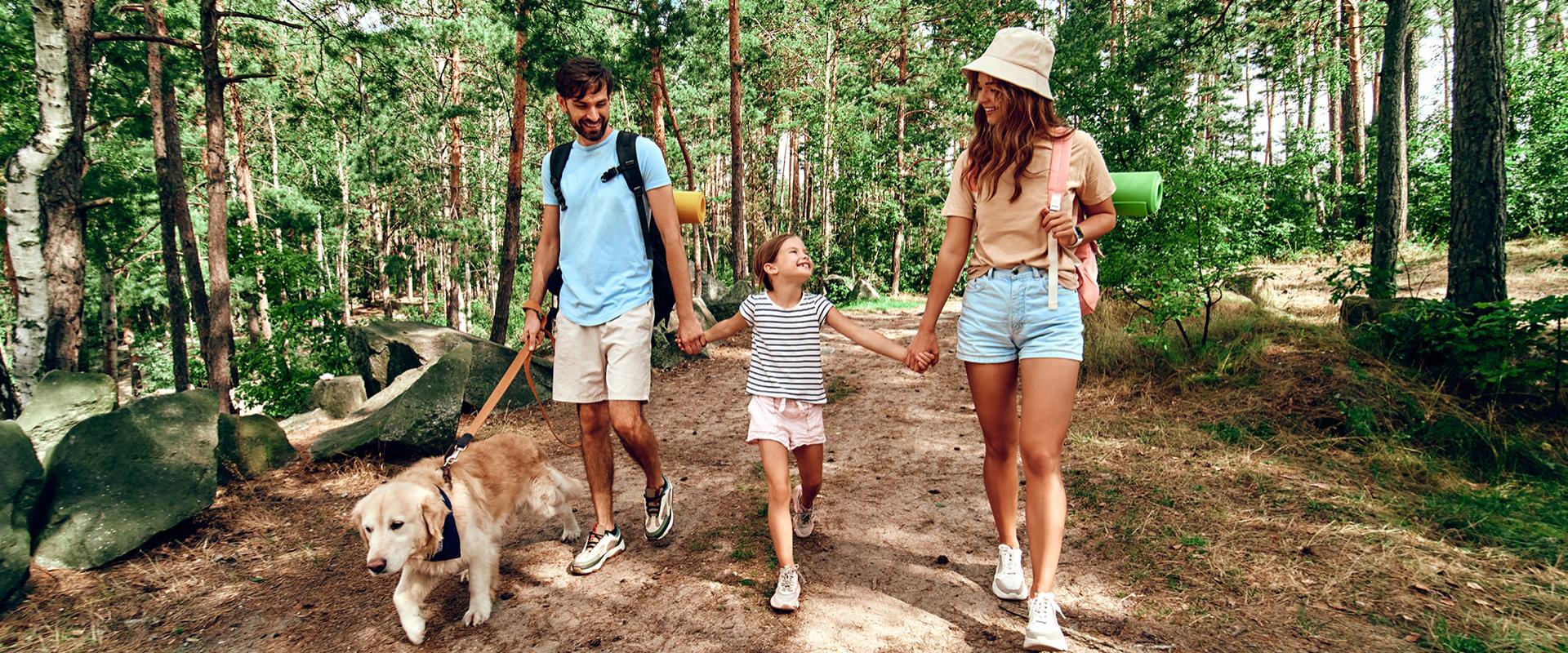 Famiglia cammina nel bosco con il cane, sorridenti e felici, durante una giornata estiva.