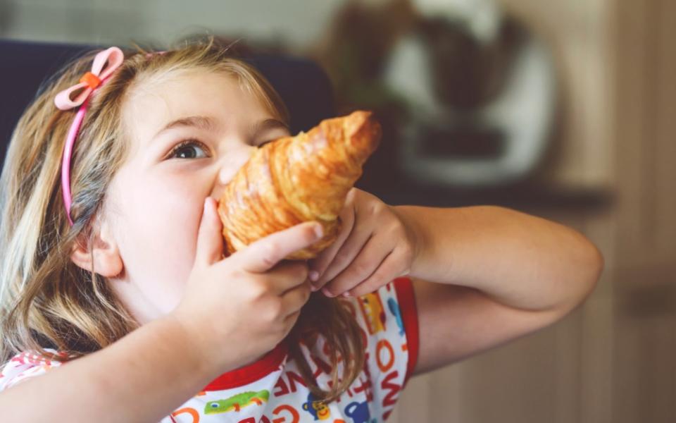 Bambina mangia un croissant con entusiasmo in cucina.