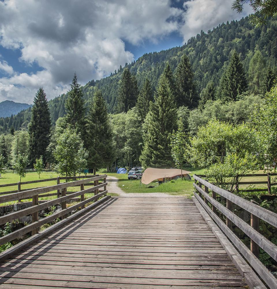 Ponte di legno in un campeggio montano, circondato da alberi e tende.